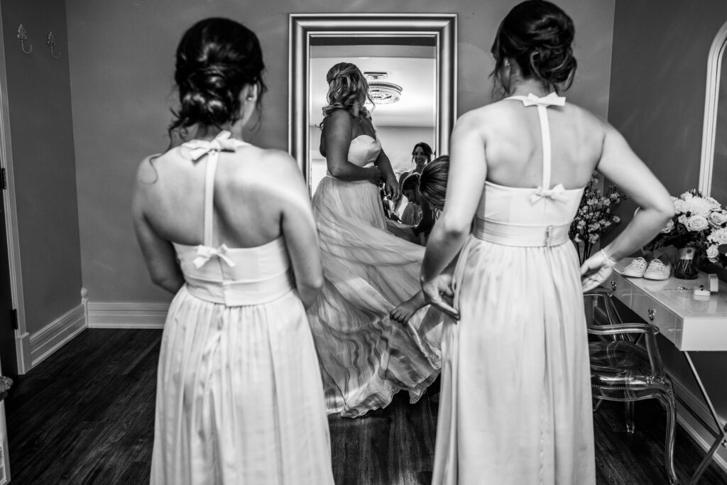 black and white photo of bridesmaids getting bride dressed looking in mirror