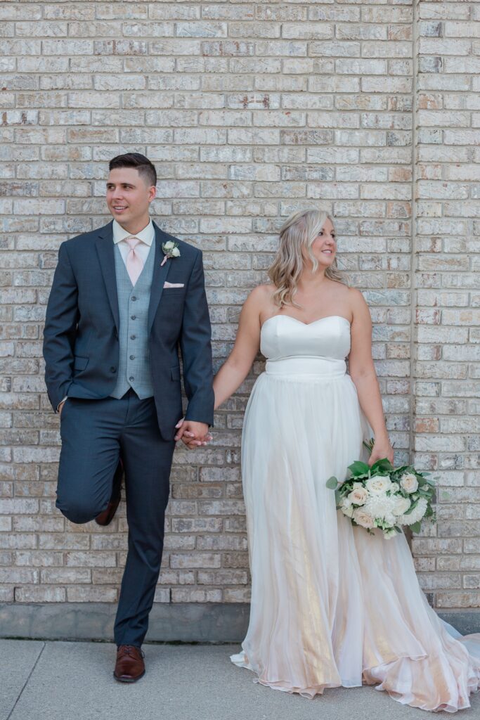 bride and groom holding hands leaning up against brick wall