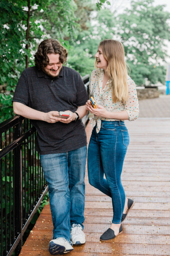Couple playing with game boys leaning along railing during their engagement session at Mill Race Park