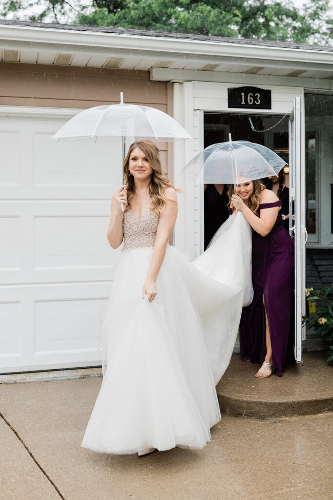 bride and her maid of honour walking out of the house into the rain on her wedding day holding clear umbrellas