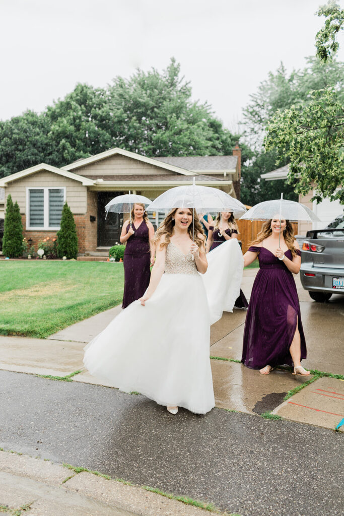 bridesmaids carrying a brides wedding gown in the rain and all holding clear umbrellas
