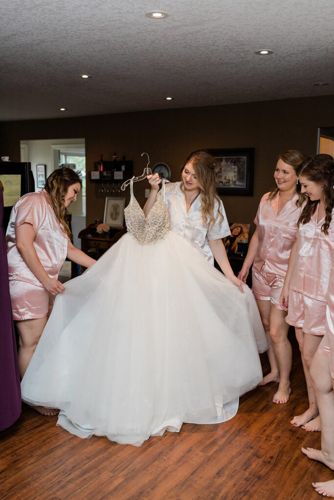 bride holding a wedding gown on a hanger with her bridesmaids standing around her in silk pj's
